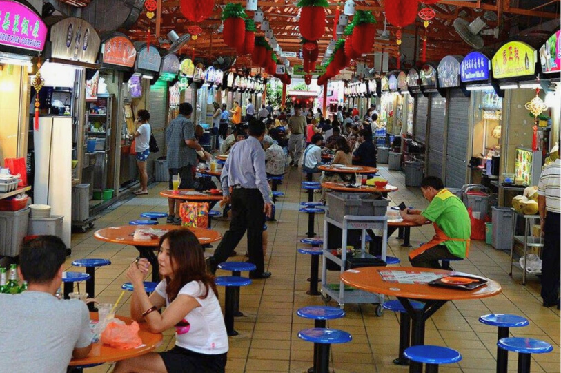 A crowded, bustling open-air hawker center in Singapore with rows of food stalls and people dining at orange circular tables.