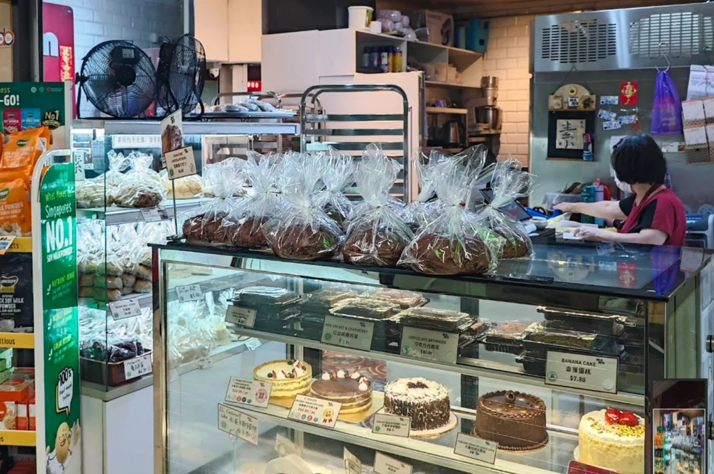 A bakery counter filled with various baked goods, including loaves of bread in clear plastic bags, chocolate brownies, and several whole cakes with frosting and fruit toppings arranged on glass shelves. A staff member is working behind the counter in the background.