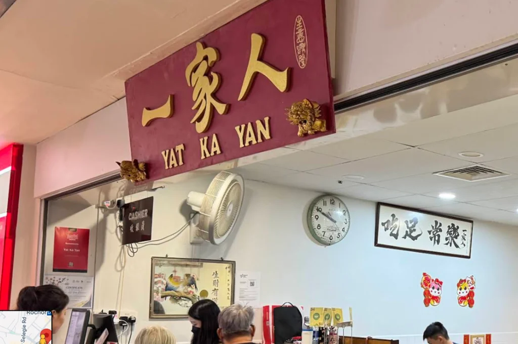 The storefront sign for "Yat Ka Yan," featuring large gold Chinese characters and the English name underneath, mounted on a deep red plaque. The interior view shows a wall with a round analog clock, a framed traditional painting, and a white sign with Chinese calligraphy, along with a staff member at the counter.