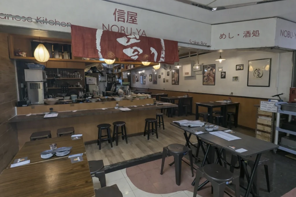 The interior of "Noru-Ya," a Japanese-style restaurant with a wooden counter, high stools, and several small tables. A large red fabric curtain (noren) hangs above the service area, and the walls are decorated with framed posters, prints, and calligraphy in a minimalist, wood-toned setting.