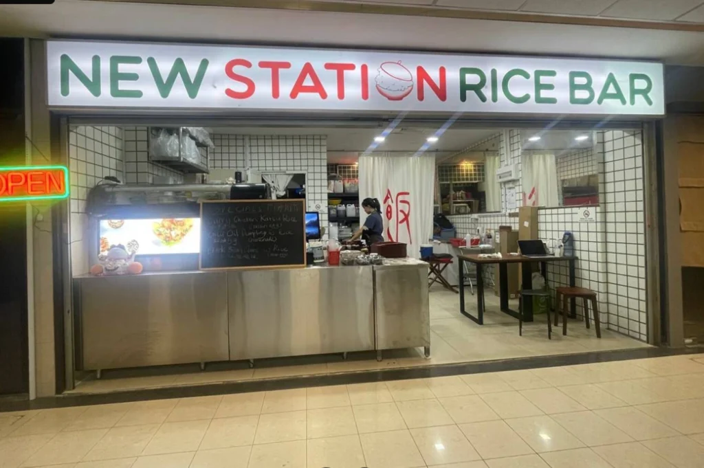 The storefront of "New Station Rice Bar," featuring a prominent illuminated sign with the name in green and red letters. The stall has a stainless steel counter, a chalkboard displaying daily specials, and white tiled walls. A staff member is visible working inside, and a glowing neon "OPEN" sign is displayed on the left.