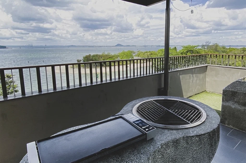 A close-up view from a balcony overlooking a scenic coastal area in Singapore, featuring a stone outdoor grill and griddle setup in the foreground, with a metal railing and a peaceful view of the sea and distant island hills under a cloudy sky.