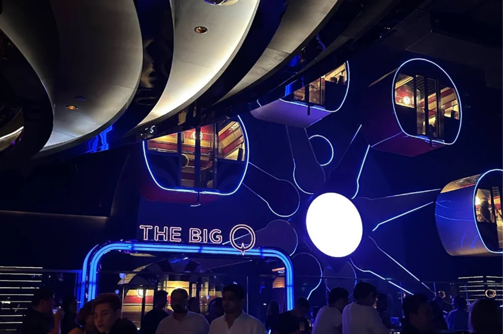 A medium shot taken inside a nightclub featuring a unique "The Big Q" sign in blue neon. The ceiling is decorated with circular, illuminated pods or private booths that look like a futuristic Ferris wheel. People are socializing in the dimly lit foreground, surrounded by blue neon accents and modern architectural curves.