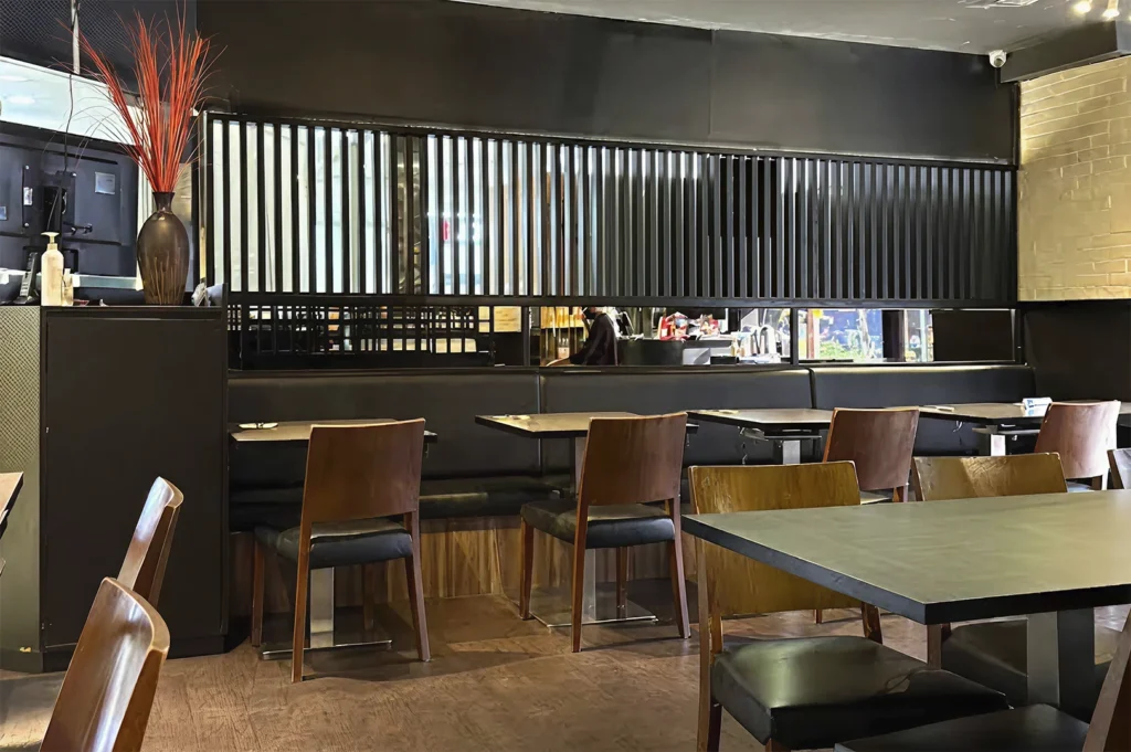 The interior of a restaurant featuring dark-toned decor, including black wall paneling, a row of booths with black leather upholstery, and wooden tables and chairs. A decorative arrangement with tall, red, grass-like stalks in a dark vase sits on a counter in the foreground.