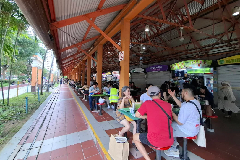 Open-air hawker center walkway, focusing on the interface between the dining area and a paved outdoor path. The scene is defined by a series of orange structural pillars supporting a corrugated metal roof, with diners seated at green tables that extend deep into the background. In the immediate foreground, a group of three people is engaged in conversation at a table, while a person in a bright neon yellow safety vest sits nearby. To the left, a red and white tiled walkway runs parallel to a lush green park area with tropical trees and a white archway in the distance. The lighting is bright and natural, capturing a typical daytime scene of social dining and urban greenery.