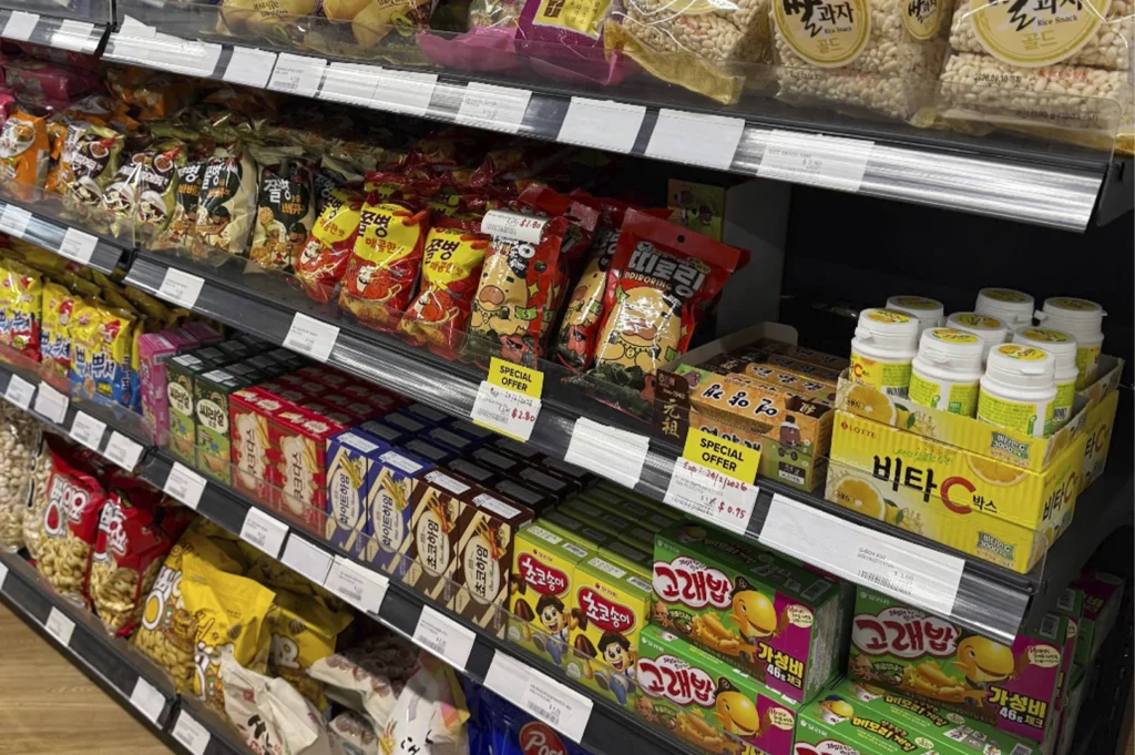 A close-up view of a retail shelf in a Korean grocery store stocked with various snack items. The shelves feature brightly colored bags of Korean snacks, including red packages of "Ddiriring" snacks, boxes of Lotte Vitamin C, and boxes of "Goraebap" (whale-shaped crackers). Many items have yellow "Special Offer" price tags attached to the shelf edge.