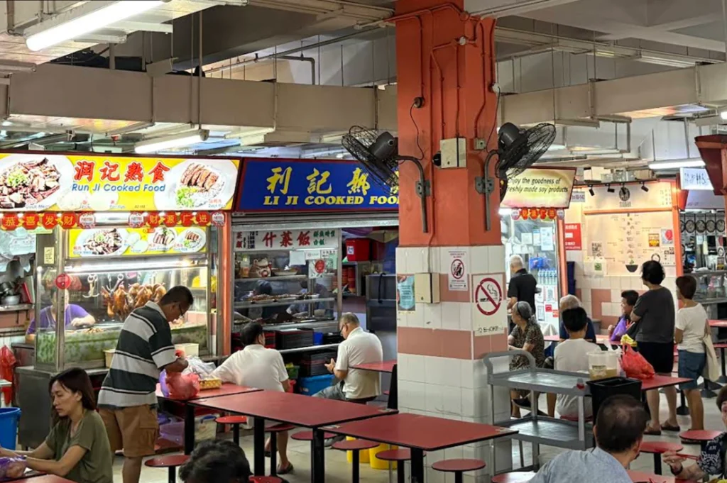 A wide-angle, documentary-style photograph of a brightly lit indoor hawker center in Singapore. In the foreground, patrons sit at communal rectangular red tables on red stools; some are eating while others look at their phones. Two prominent food stalls are visible in the background: "Run Ji Cooked Food" on the left, displaying roasted ducks in a glass case, and "Li Ji Cooked Food" next to it with a blue sign. A large square concrete pillar painted orange and white stands in the center of the frame, equipped with two black industrial oscillating fans and "No Smoking" signs. The ceiling is high with exposed pipes and industrial light fixtures, reflecting a functional and authentic local dining environment. The overall lighting is natural and even, highlighting the everyday textures of the food court.