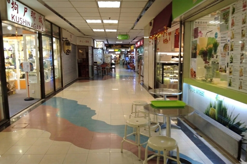 An indoor view of a brightly lit, quiet shopping mall corridor with white tiled floors. Small food stalls and shops line both sides, including "Kushinagara." In the foreground, a simple round metal table and four white stools sit outside a juice stall.