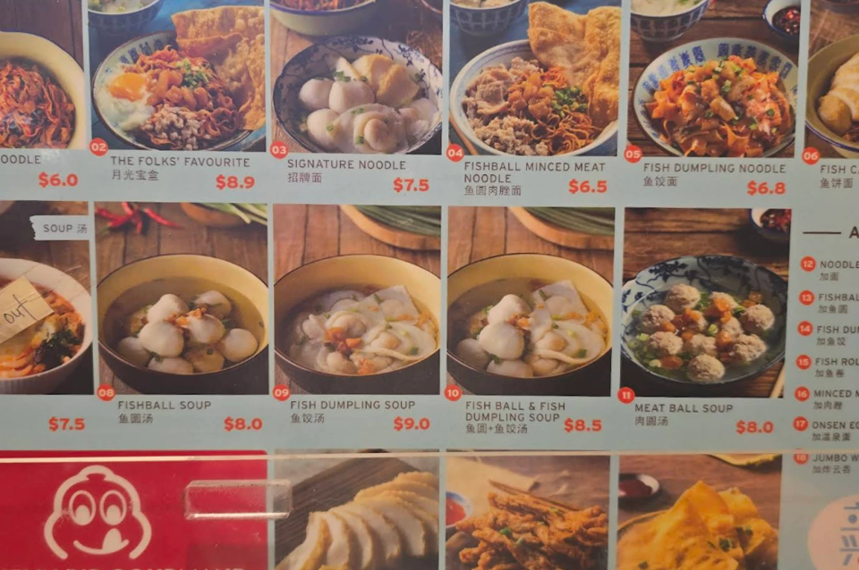 Colorful menu board from a Singaporean hawker stall, likely specialized in fishball noodles. The menu is organized into a grid with high-resolution photos of each dish, accompanied by names in English and Chinese, and prices in red. The top row features dry and soup noodle options like "The Folks' Favourite" for $8.90 and "Signature Noodle" for $7.50, which includes fishballs and wontons. The middle row focuses on soup-based dishes, such as "Fishball Soup" for $8.00 and "Meat Ball Soup" for $8.00, served in traditional patterned ceramic bowls. A side panel on the right lists add-ons like extra noodles, fishballs, and onsen eggs. At the bottom left, a portion of a red logo featuring a smiling cartoon face is visible. The lighting is warm and clear, emphasizing the textures of the food.