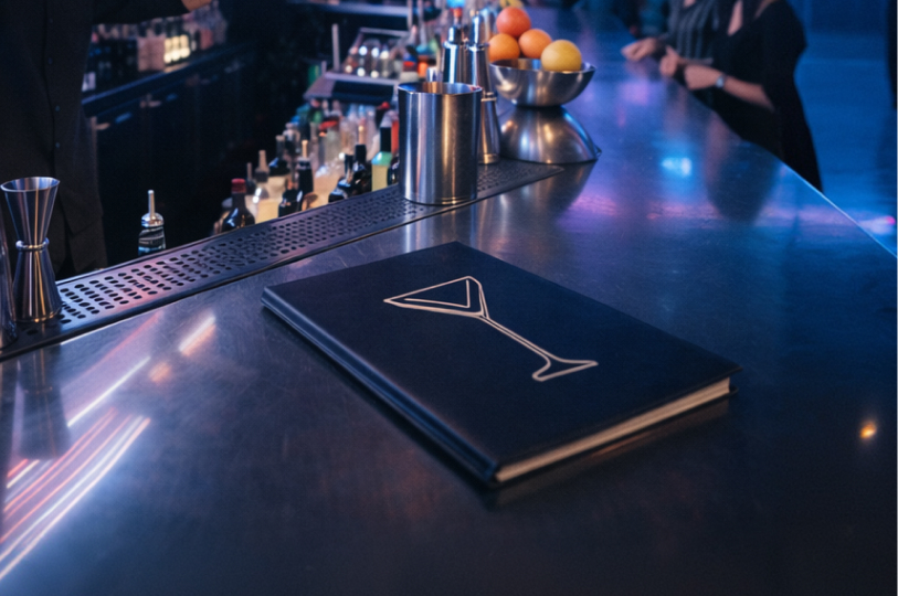 A sleek black cocktail menu with a white martini glass icon lying on a polished bar counter next to bartending tools.