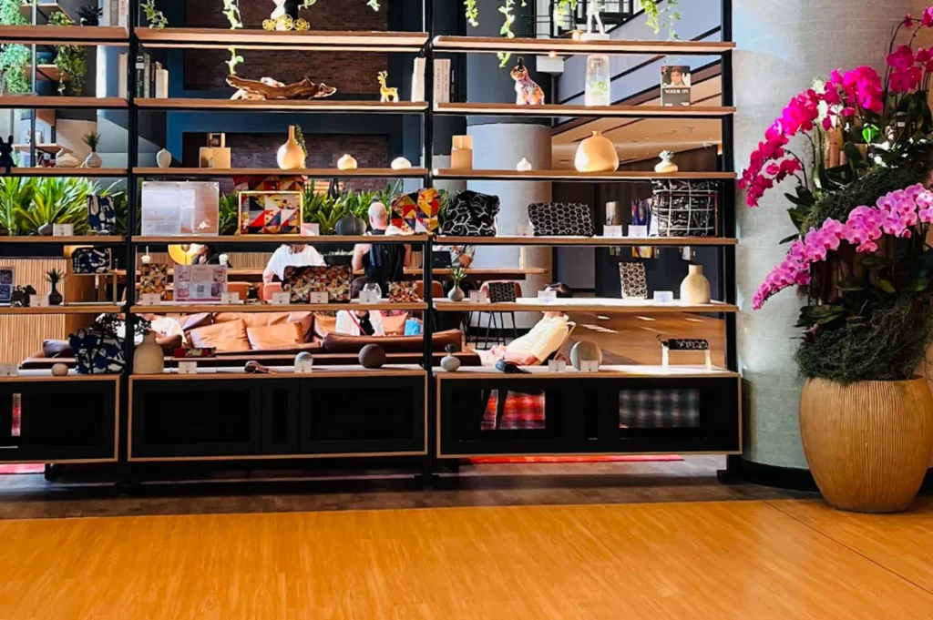 A wide, eye-level shot of a contemporary hotel lobby featuring a large, floor-to-ceiling black metal shelving unit. The shelves are sparsely decorated with minimalist vases, books, and colorful geometric-patterned bags. Behind the shelving, a lounge area is visible with guests seated on low leather sofas. To the right, a massive terracotta pot holds a vibrant arrangement of bright pink orchids, adding a splash of color against the warm wooden floors and dark interior accents.