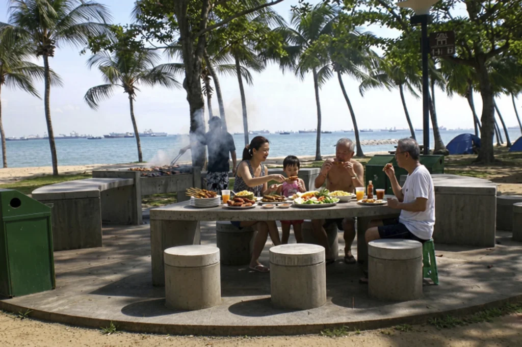 A family of four enjoys an outdoor BBQ feast at a concrete picnic table near the coast. Behind them, palm trees line a sandy beach, and several large ships are visible on the calm blue sea under a clear sky.