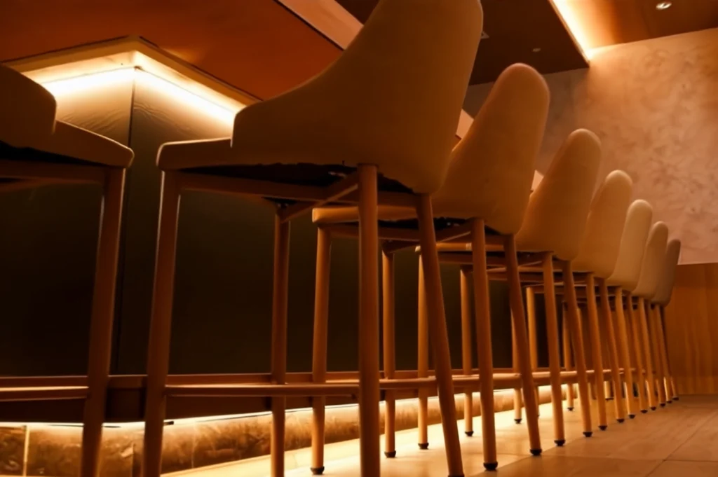 A low-angle, atmospheric shot of a row of modern, cream-colored bar stools lined up against a dark dining counter. Warm, indirect amber lighting glows from underneath the counter and along the ceiling cove, casting soft shadows. The minimalist interior design and warm tones create a sophisticated, intimate dining ambiance.
