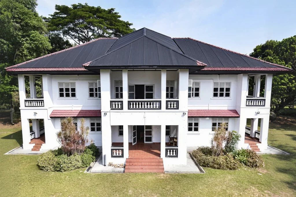 An architectural exterior shot of a colonial-style white bungalow in Singapore, featuring a symmetrical design with a dark gray hip roof, a central balcony with ornate white balusters, and mature trees in the surrounding lawn area.