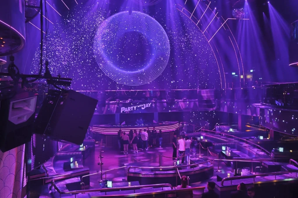 An eye-level, panoramic view of a nightclub dance floor from a slightly elevated balcony. The room is bathed in dramatic purple lighting, highlighting a large circular art installation or screen on the back wall. The space is sparsely populated, with a DJ booth marked "Party with Jay" in the center and sleek, curved seating areas arranged around the dance floor.
