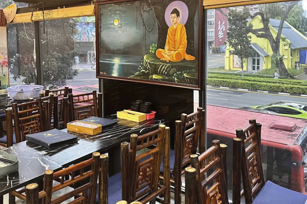 A dining area inside a restaurant featuring a large, artistic painting of a monk meditating under a tree hanging above a black marble table. The space is furnished with dark wood chairs featuring intricate cut-out patterns, and a large window looks out onto a street with a yellow building in the distance.