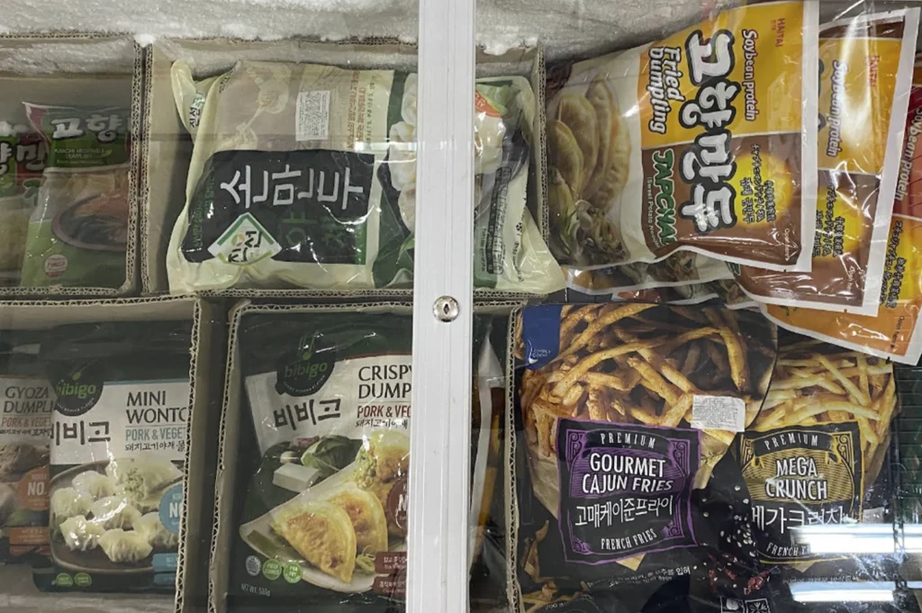 A view inside a chest freezer at a Korean grocery store. The freezer contains various frozen food items in bags, including Bibigo brand dumplings (gyoza, mini wontons, and pork and vegetable dumplings), other varieties of Korean dumplings, and bags of premium frozen french fries labeled "Gourmet Cajun Fries" and "Mega Crunch."