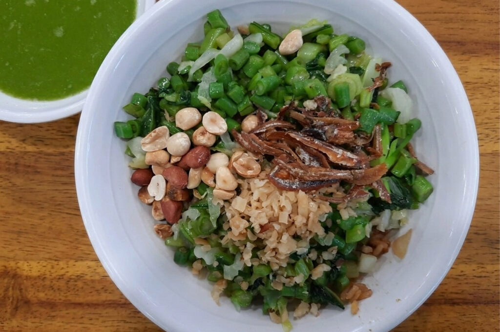 A top-down view of a Thunder Tea Rice (Lei Cha) bowl. The white bowl is filled with brown rice topped with chopped green beans, peanuts, dried anchovies, and preserved radish. A small side bowl contains the signature vibrant green herbal tea soup.