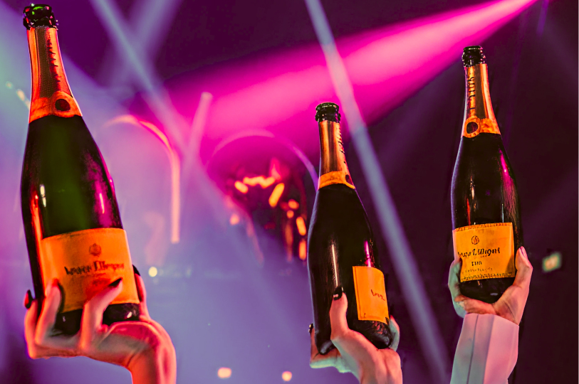 Three hands holding up bottles of Veuve Clicquot champagne under vibrant pink and purple club lights.