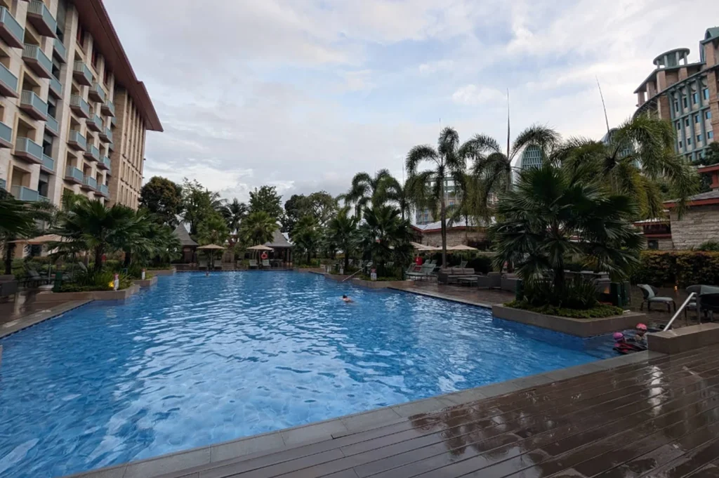 A wide landscape shot of a large, rectangular outdoor swimming pool under a cloudy, overcast sky. The blue water is slightly ruffled, with a few guests swimming. The pool is surrounded by a wet wooden deck, reflecting the muted light. Mature palm trees and tropical shrubbery line the perimeter, separating the pool from the hotel buildings. In the distance, the unique tiered architecture of Resorts World Sentosa structures can be seen against the horizon.