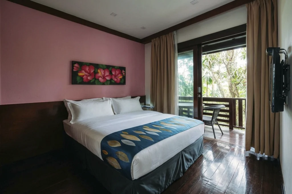 The interior of a cozy chalet bedroom featuring a large bed with white linens and a blue decorative runner. The room has a pink accent wall adorned with a painting of hibiscus flowers and a sliding glass door that opens onto a private wooden balcony surrounded by lush greenery.