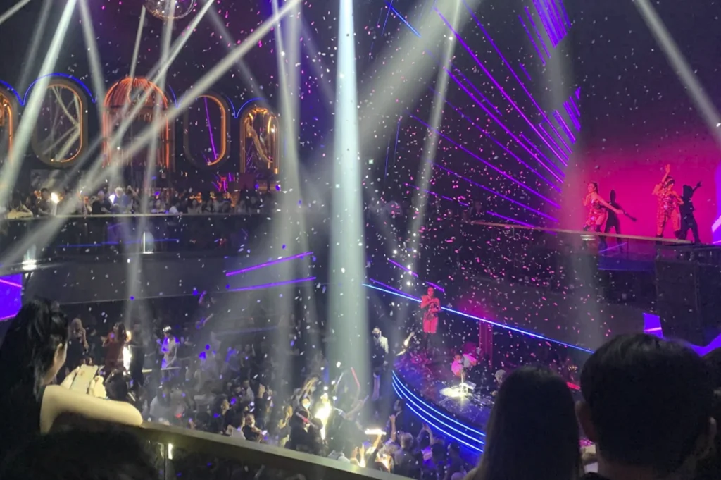 An action shot from a balcony overlooking a crowded, energetic dance floor. White confetti is raining down on the club-goers as bright white spotlights beam from above. On a raised platform to the right, performers are dancing in front of a bright pink screen, adding to the high-energy party atmosphere.