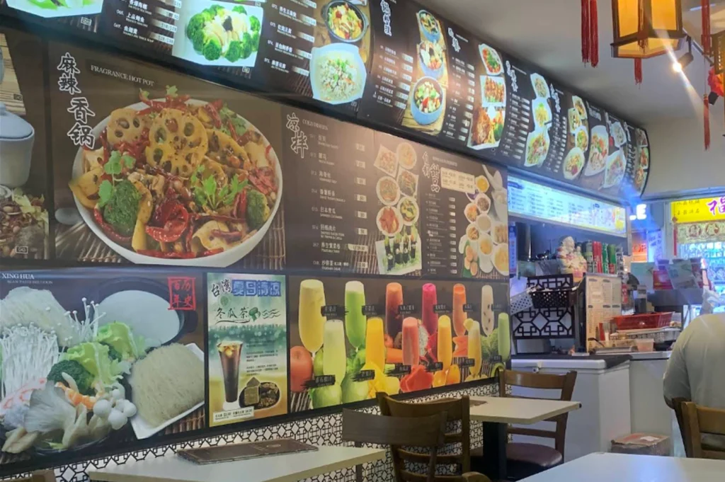 A wall-mounted menu board in a restaurant showcasing various food dishes with large photographs, including a bowl of "Fragrance Hot Pot" filled with colorful vegetables, lotus root, and spices, alongside other menu items and a selection of colorful fruit drink images.