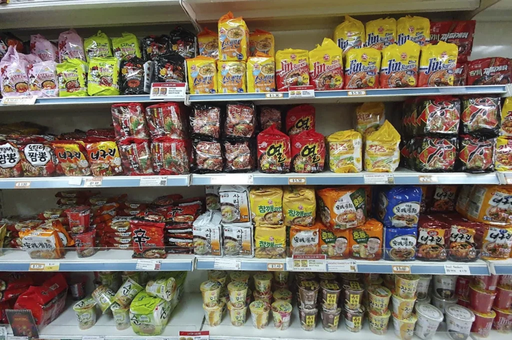 A well-stocked shelf of instant noodles at a Korean grocery store. Rows of colorful packets of Korean ramyeon are neatly arranged. Brands visible include Jin Ramen, Neoguri, and various spicy flavor varieties. Multiple yellow price tags are attached to the shelves, and some noodle cups are stacked on the bottom shelf.