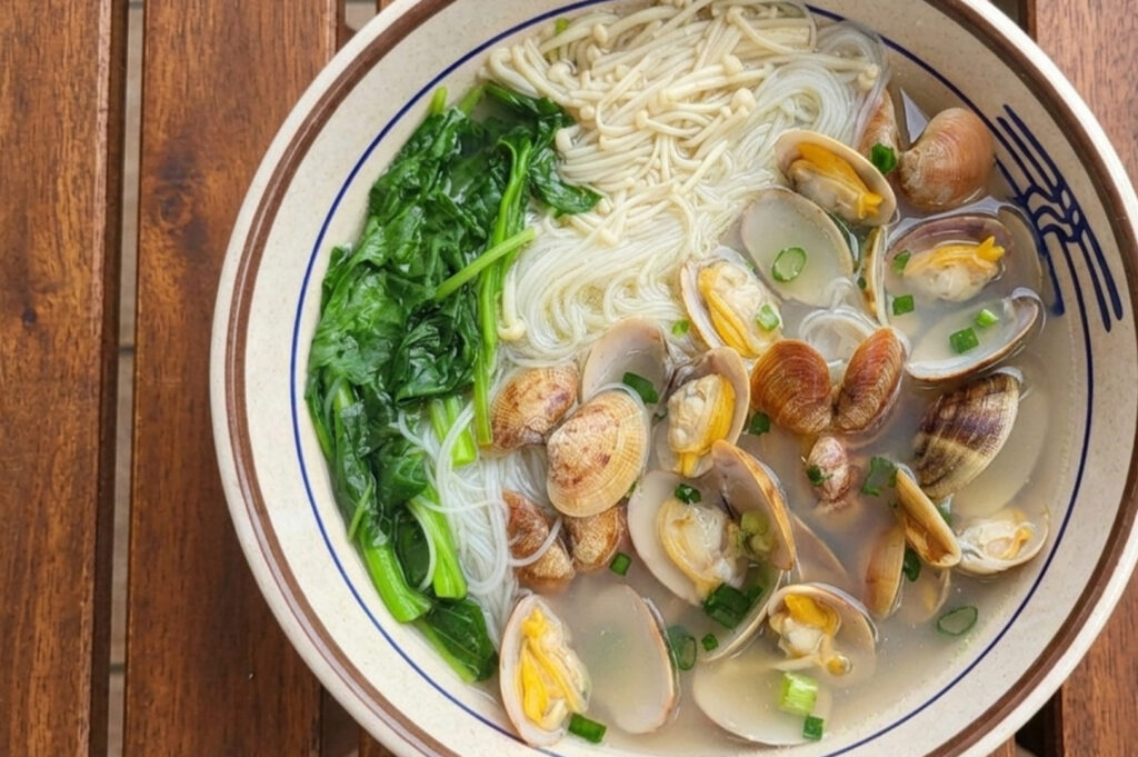 A bowl of Clam Bee Hoon soup served on a wooden table. The clear broth contains thin rice vermicelli, numerous open clams, enoki mushrooms, bright green leafy vegetables, and a garnish of chopped spring onions.