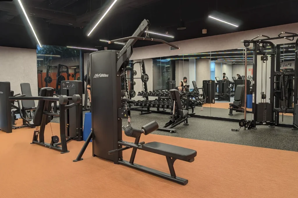 A bright, clean interior shot of a modern gym. The room features orange-toned rubber flooring and a variety of professional Life Fitness strength-training equipment, including a cable lat pulldown machine in the foreground and a rack of dumbbells in the back. Large floor-to-ceiling mirrors line the far wall, reflecting the equipment and a few people working out. The ceiling is black with recessed linear LED lighting strips providing a sharp, industrial feel.