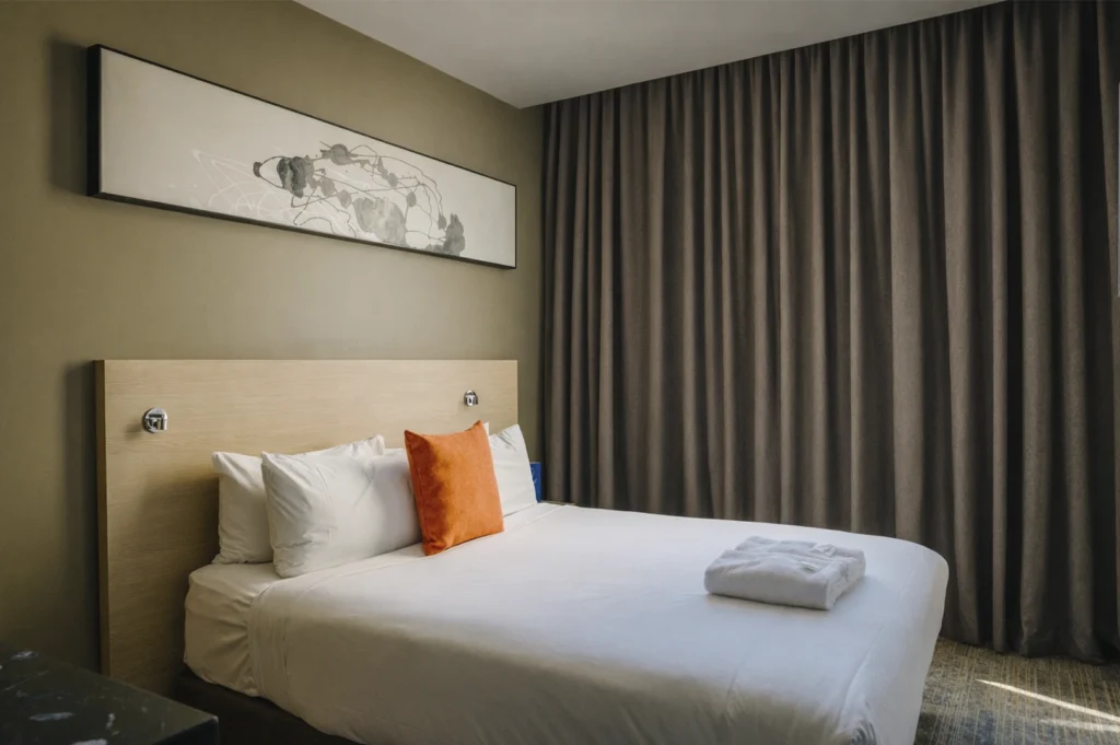 A cozy, modern hotel bedroom interior in Singapore featuring a comfortable bed with white linens and a bright orange accent pillow, a light wood headboard, brown curtains, and a piece of abstract art hanging on a taupe-colored wall.