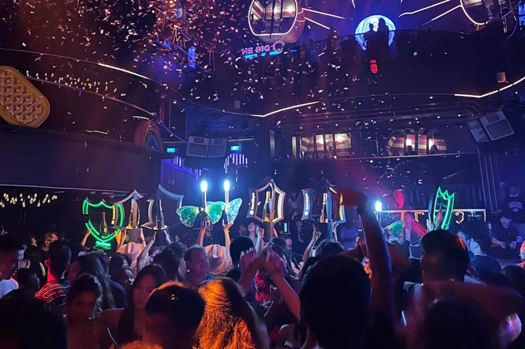 A low-angle, immersive shot from the middle of a packed dance floor. People are cheering and dancing, with several guests holding up glowing, branded champagne bottle displays. Confetti fills the air, and the room is illuminated by a mix of blue, purple, and orange lighting, conveying a sense of celebration and excitement.