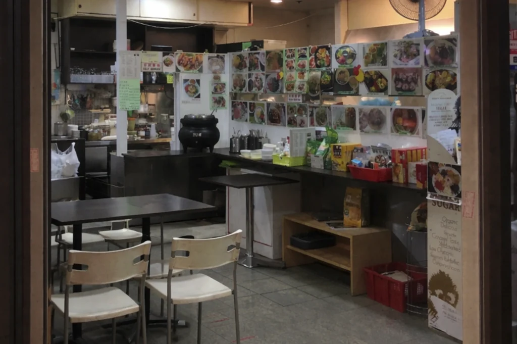 A casual, open-concept kitchen area inside a food establishment, with a central black cooking pot, wooden tables for seating, and numerous small photos of different dishes displayed on the back wall above a service counter.
