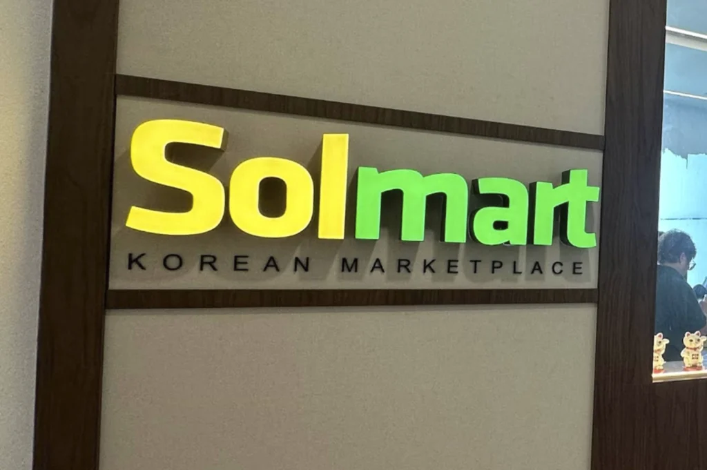 A close-up shot of the "Solmart" store sign, featuring the name in large, 3D-style yellow and green lettering mounted on a light-colored panel with a wooden frame. "KOREAN MARKETPLACE" is printed in black lettering beneath the main logo. To the right, through a glass section, a small portion of the store interior and a decorative cat figurine are visible.