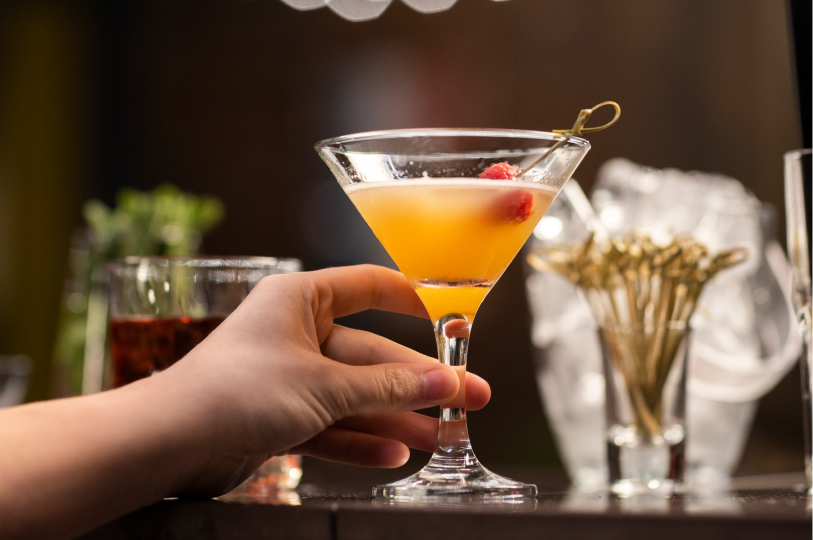 A hand reaching for a yellow martini garnished with a raspberry, sitting on a bar with ice buckets and toothpicks in the background.