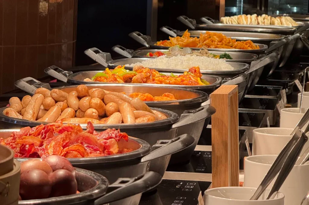 A close-up, shallow-depth-of-field shot of a breakfast buffet line. Large stainless steel chafing dishes are filled with various hot foods, including crispy bacon in the front, followed by a tray of breakfast sausages, golden-brown stir-fried noodles, steamed white rice, and other savory dishes extending into the background. The lighting is warm and focused on the food, highlighting the textures of the ingredients.