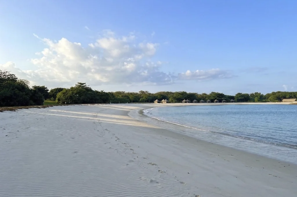 A peaceful, wide-angle shot of a pristine white sand beach curving along clear turquoise water. The shoreline is bordered by dense green trees, and several small thatched-roof huts are nestled in the distance under a bright blue sky.