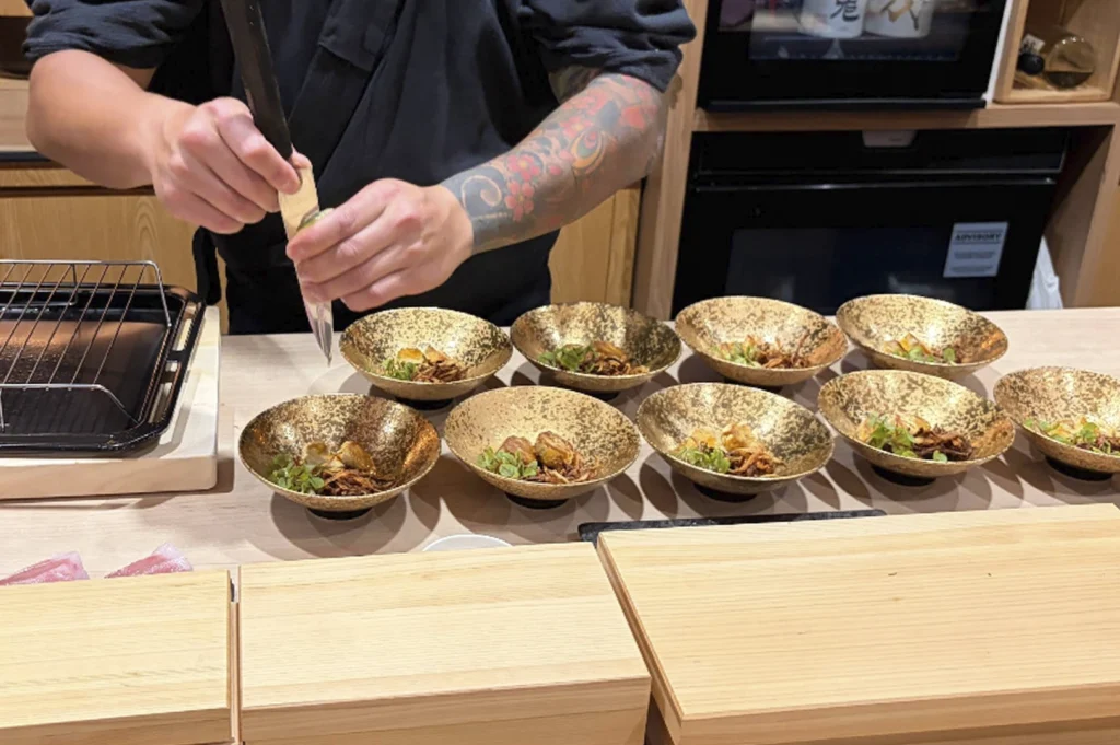 An action shot of a chef's hands as he finishes a series of small dishes. He is squeezing a lime over eight ornate, textured gold bowls arranged in two rows on a light wood counter. Each bowl contains a small, elegantly plated appetizer with microgreens and crispy elements. The chef wears a dark uniform, and his forearm features a colorful, intricate floral tattoo.