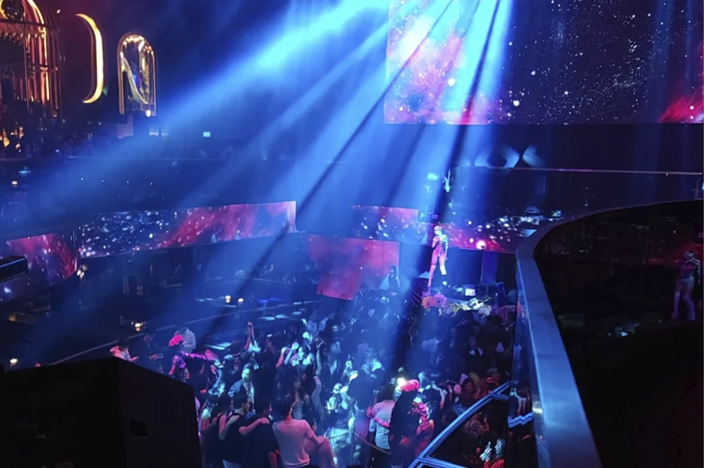 An elevated, high-angle view looking down at a crowded dance floor. Strong, focused blue beams of light cut through the dark air, illuminating the dancers below. A large screen behind the performers displays a vibrant, space-themed nebula graphic. A performer is visible on a small stage in the center of the floor.