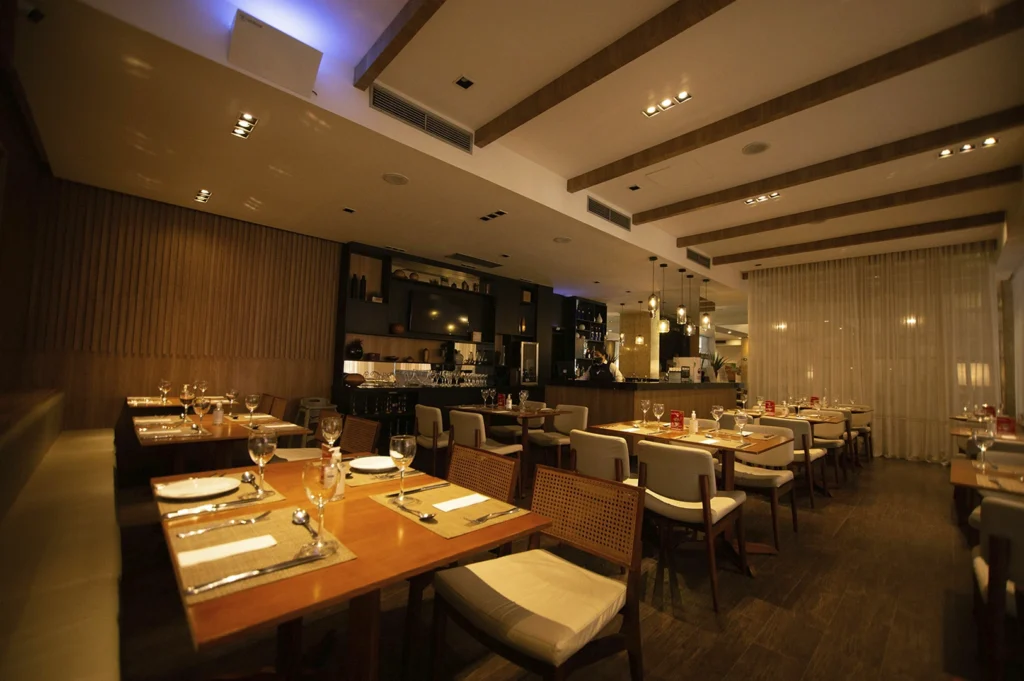 A wide-angle shot of a modern, upscale restaurant interior. The space features warm wooden wall paneling, clean-lined furniture with wicker-back chairs, and polished wood floors. The tables are meticulously set with white plates and glassware. Soft, recessed ceiling lights and decorative glass pendant lamps over a dark bar area create a sophisticated, inviting evening ambiance.