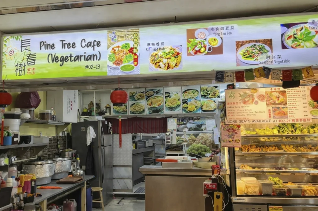 The storefront of "Pine Tree Cafe (Vegetarian)," featuring a brightly lit overhead sign with the name, Chinese characters, and images of various vegetarian dishes. The stall includes a service counter, a display case filled with fresh produce and prepared ingredients, and red lanterns hanging from the ceiling.