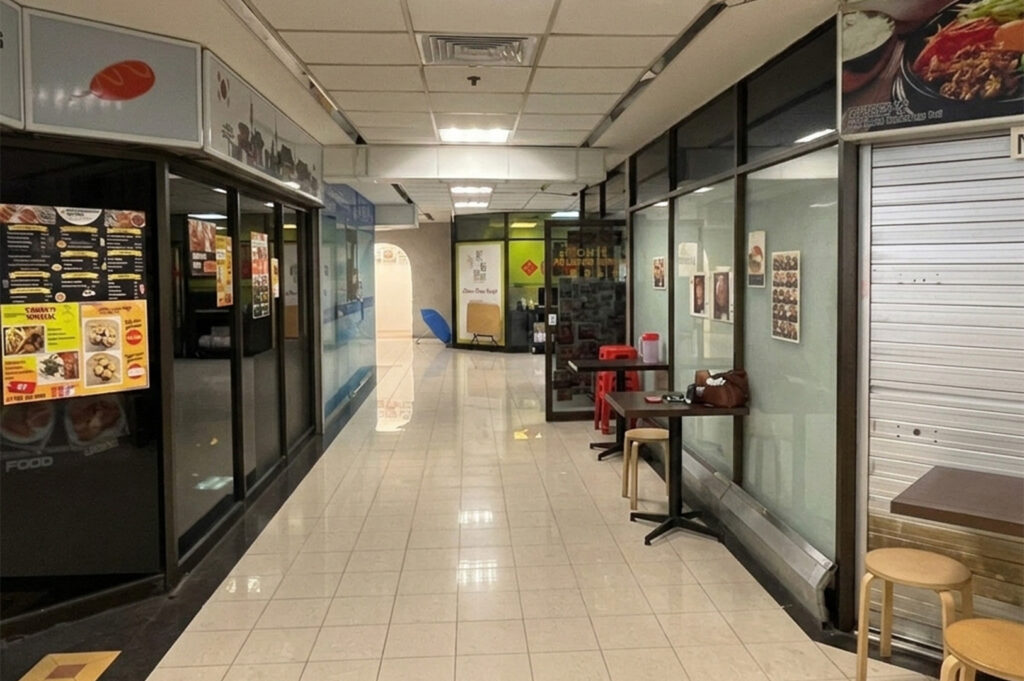 A perspective view of a narrow, sparsely populated mall hallway with white ceiling tiles and fluorescent lighting. Small eateries with glass storefronts and menus line the path, with a few simple wooden tables and stools placed in the walkway for diners.