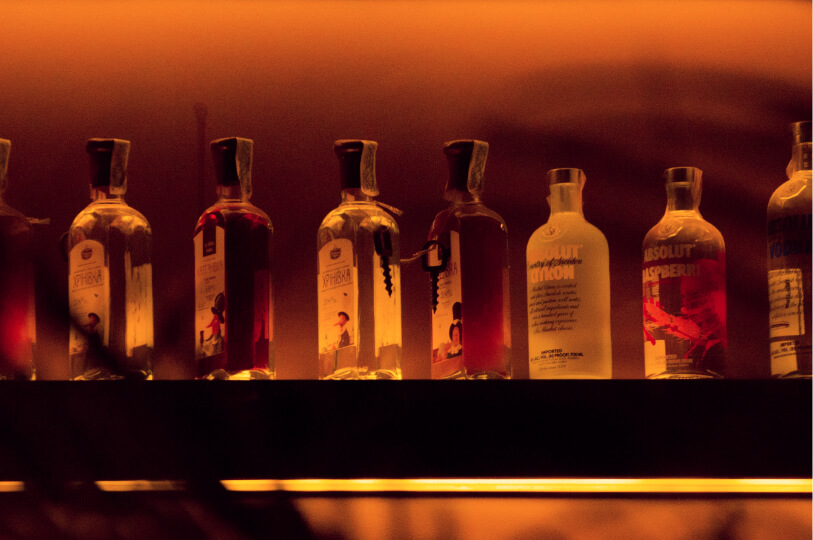 A row of vodka bottles, including Absolut Citron and Raspberry, backlit by a warm, glowing orange light on a bar shelf.