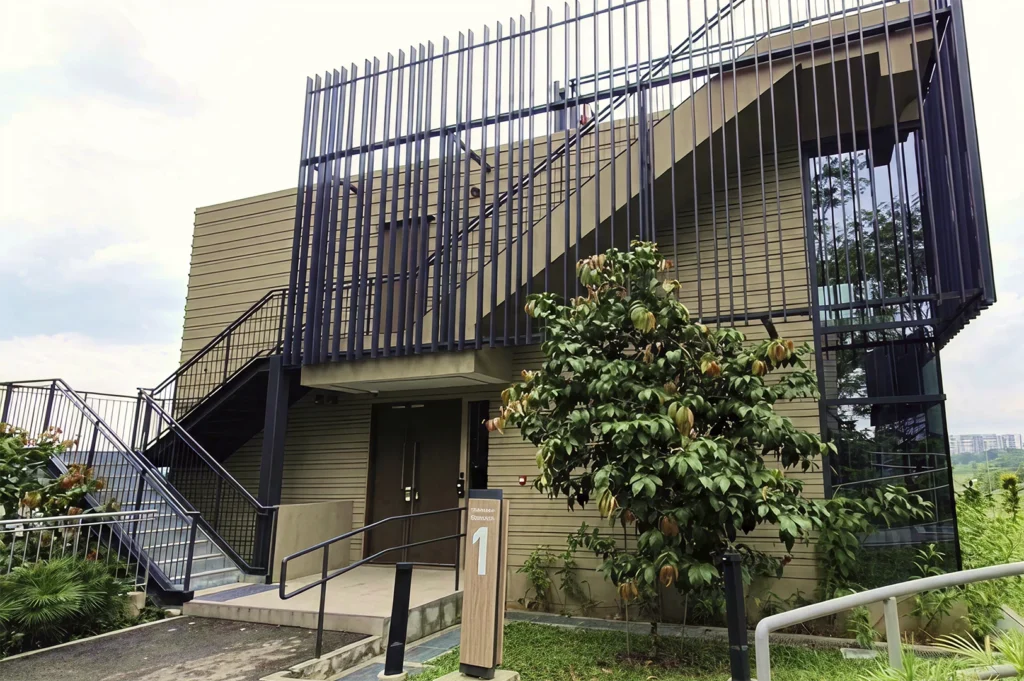 A contemporary two-story chalet building with olive-green horizontal siding and a prominent black metal staircase on the exterior. Large glass windows reflect the surrounding trees, and a sign with the number "1" stands at the entrance.