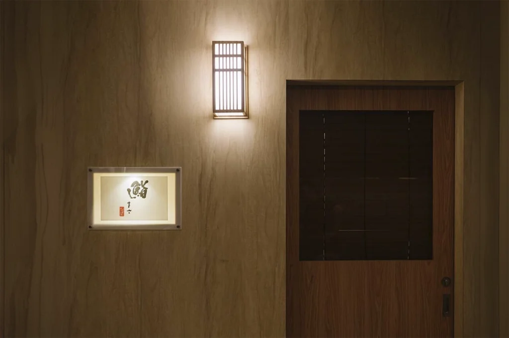 A minimalist shot of a restaurant entrance featuring a light-toned wood-paneled wall. To the left, a small, glowing rectangular sign displays Japanese calligraphy and a red seal. Above the wooden door, a rectangular lantern with a vertical slat design casts a warm, downward glow. The overall aesthetic is clean, traditional, and understated.