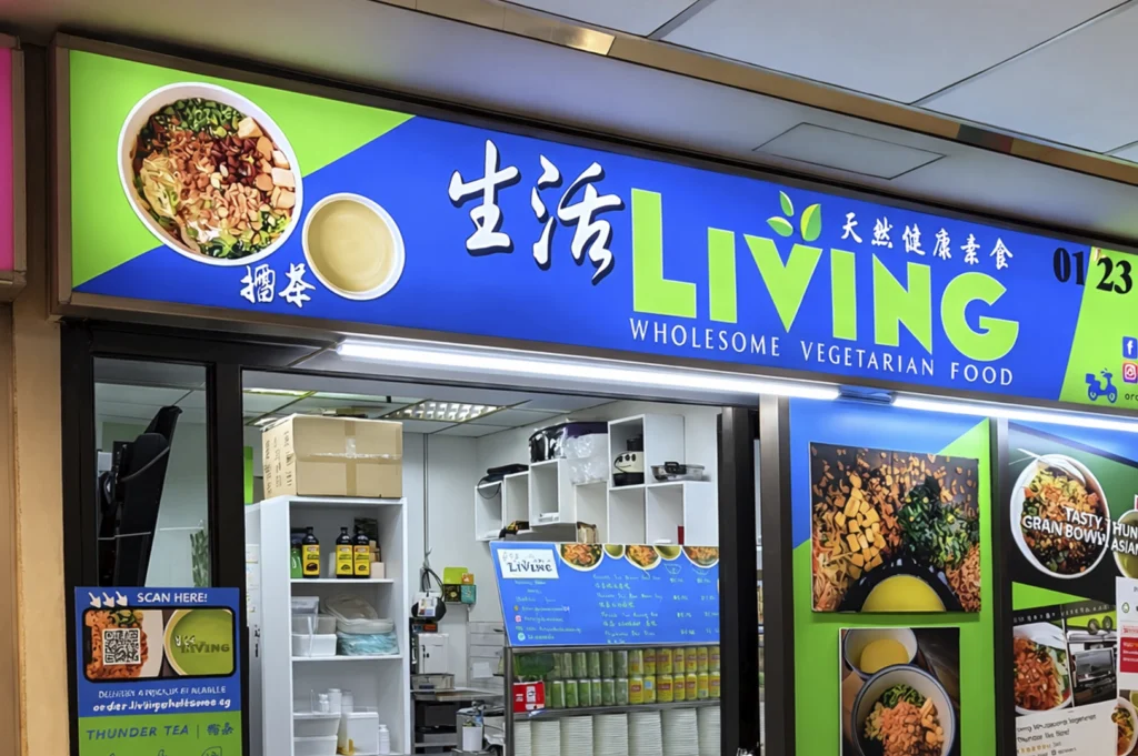 The storefront of "Living Wholesome Vegetarian Food," featuring a vibrant blue and green sign with the name in large lettering, Chinese characters, and a photo of a bowl of food. The entrance displays a QR code scanner and photographs of menu items, including a noodle bowl.