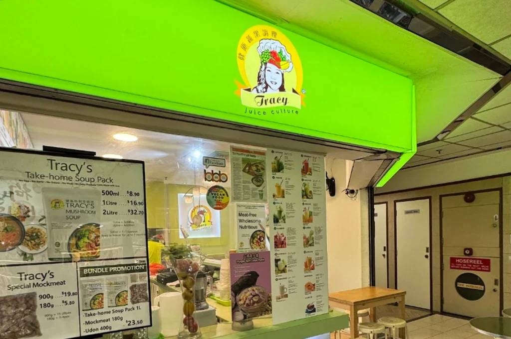 The storefront of "Tracy Juice Culture," with a bright green sign featuring a cartoon logo of a person wearing a chef's hat. The stall displays promotional posters for take-home soup packs, vegetarian mock meats, and a menu list on a transparent partition, with a small seating area nearby.
