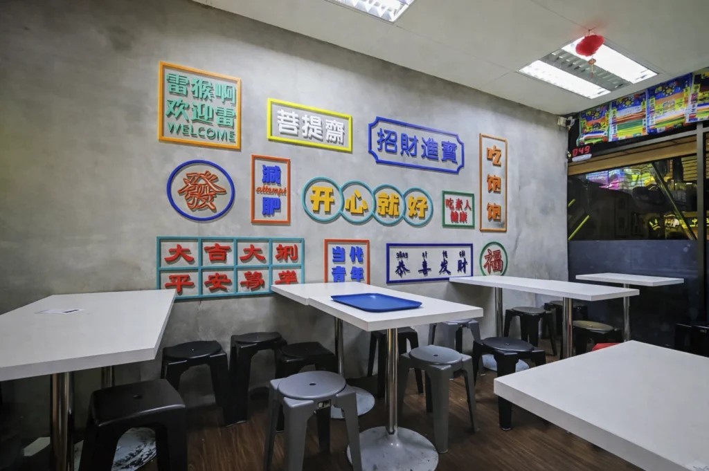 A dining area featuring a light grey wall decorated with numerous colorful, framed signs containing Chinese characters, including phrases like "WELCOME," "Happy," and "Fortune." The room is furnished with simple white tables and grey and black stools.