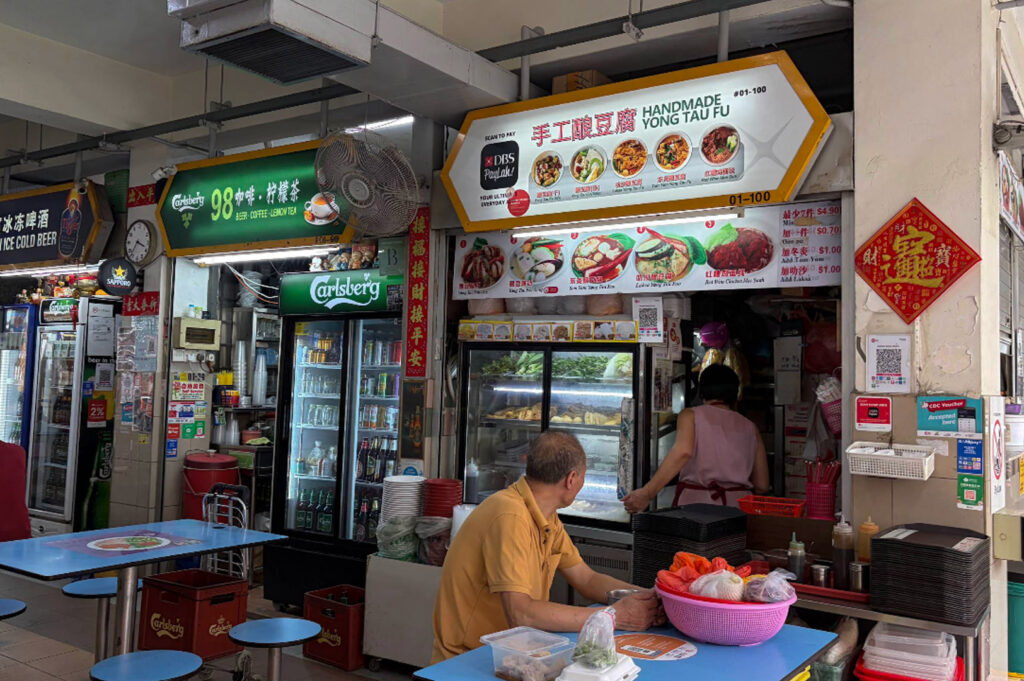A vibrant shot of a Singaporean hawker center stall named "Handmade Yong Tau Fu." The stall features a bright yellow hexagonal sign with photos of various noodle and tofu dishes. A man in a yellow shirt sits at a blue table in the foreground, while a female worker is visible inside the stall. To the left, another stall sells coffee and beer, with green Carlsberg branding and refrigerators stocked with drinks.