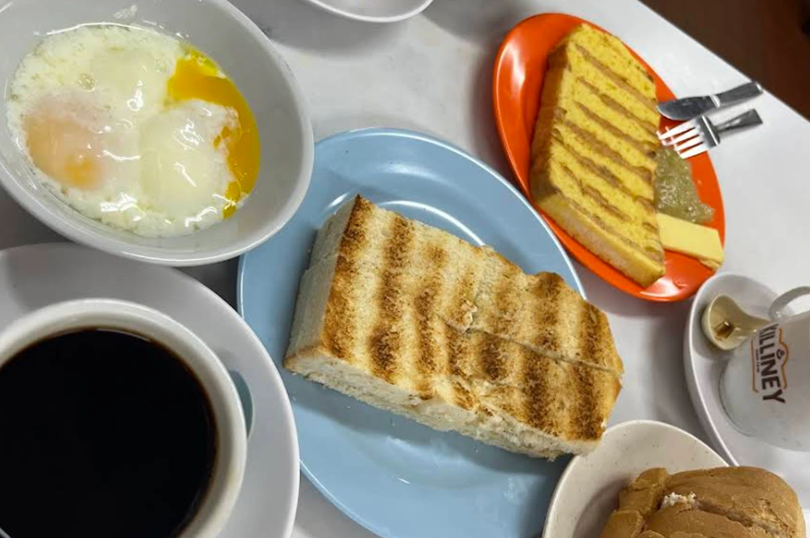A classic Singaporean breakfast spread served on a white tabletop. The meal includes two soft-boiled eggs in a white porcelain bowl with runny yolks, a thick slice of golden-brown toasted bread on a light blue plate, and another plate of French toast on a bright orange dish served with a side of kaya jam and butter. A cup of dark, rich black coffee (Kopi O) is visible in the lower-left corner, and a traditional white ceramic mug with the red "Killiney" logo sits to the right. The lighting is bright and natural, highlighting the textures of the charred toast and the glossy eggs.